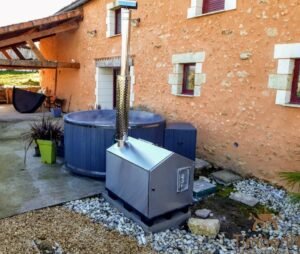 Rundes TimberIN Nordic External Comfort Badefass mit blauer Außenverkleidung vor einem Steinhaus, verbunden mit einem großen externen Pellet-Heizgerät und hohem Edelstahl-Schornstein