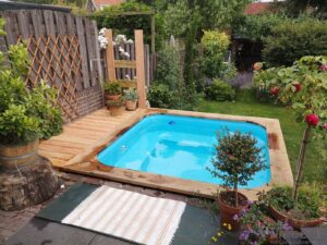 Quadratisches TimberIN Nordic Square Badefass, mit Sichtschutz aus Holz oder Stein, umgeben von Gartenbepflanzung, auf einer Holzterrasse, in einem angelegten Garten