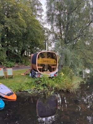 Rückansicht mit Panoramaverglasung der mobilen Sauna TimberIN Nordic Square auf einem Anhänger an einem See