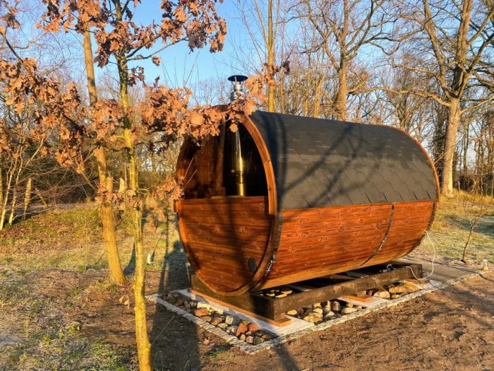 Fasssauna mit dunklem Dach und Holzunterbau zwischen Bäumen