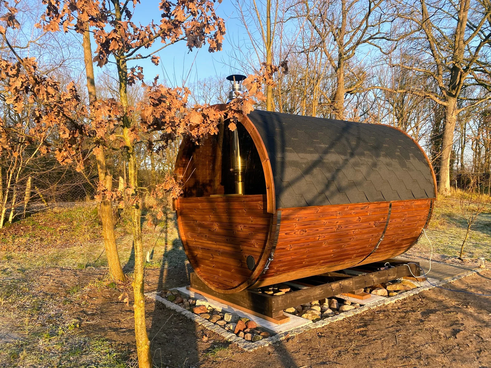 Fasssauna mit dunklem Dach und Holzunterbau zwischen Bäumen