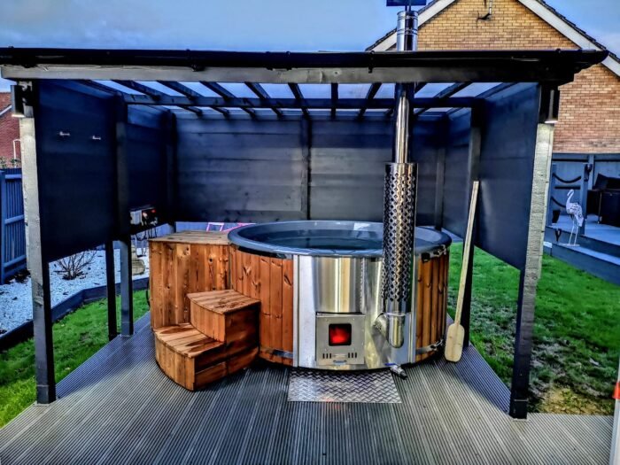 Main subcategory: Badefass mit Holzofen 12 alt="Badefass mit integriertem Holzofen aus Edelstahl – moderner Badezuber mit Holzofen unter Überdachung"