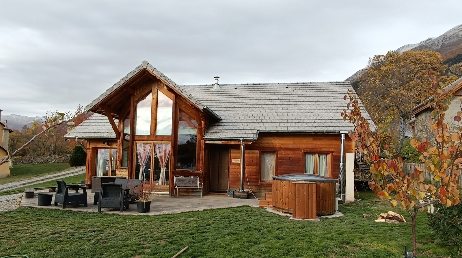 kosten 1 Rundes Holzbadefass mit dunkler isolierter Abdeckung und Außenofen neben der Holzterrasse eines Chalets mit Herbstbäumen und Bergen im Hintergrund