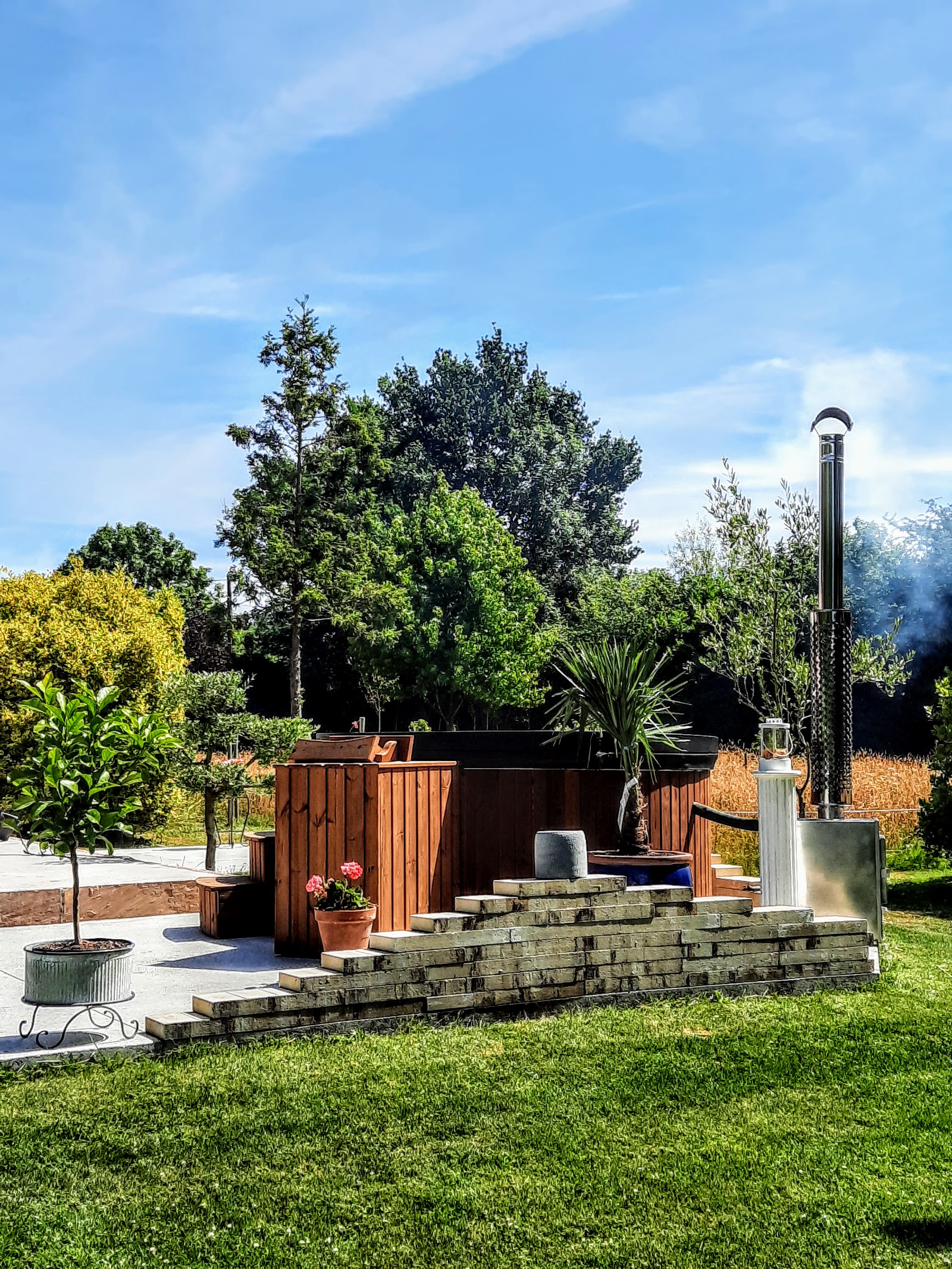 Badefass vs Pool 1 Rundes Holzbadefass mit Außenholzofen und Edelstahlschornstein auf einer Terrasse im Garten mit Rasen, Kübelpflanzen und Steinstufen