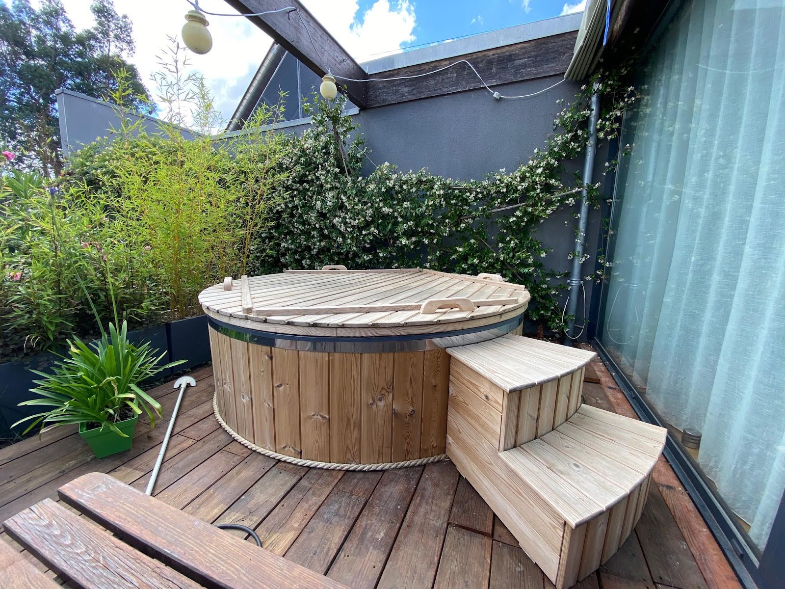 Rundes Holz-Badefass mit Holzofen auf einem Holzdeck in einem kleinen Innenhof am Haus
