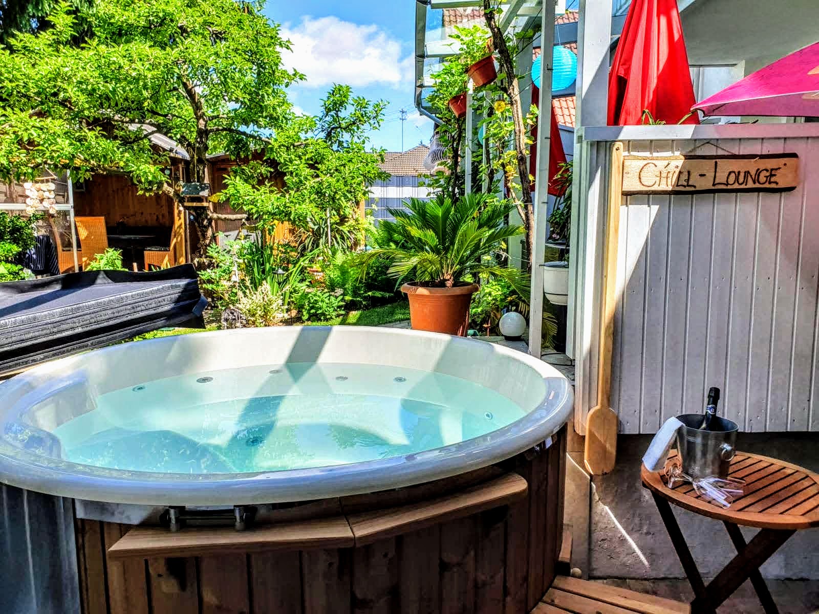 Temperatur 1 Rundes Badefass mit weißer Innenwanne und Holzverkleidung auf einer Terrasse mit Pergola, Sonnenschirmen, Kübelpflanzen und einem Holzschild