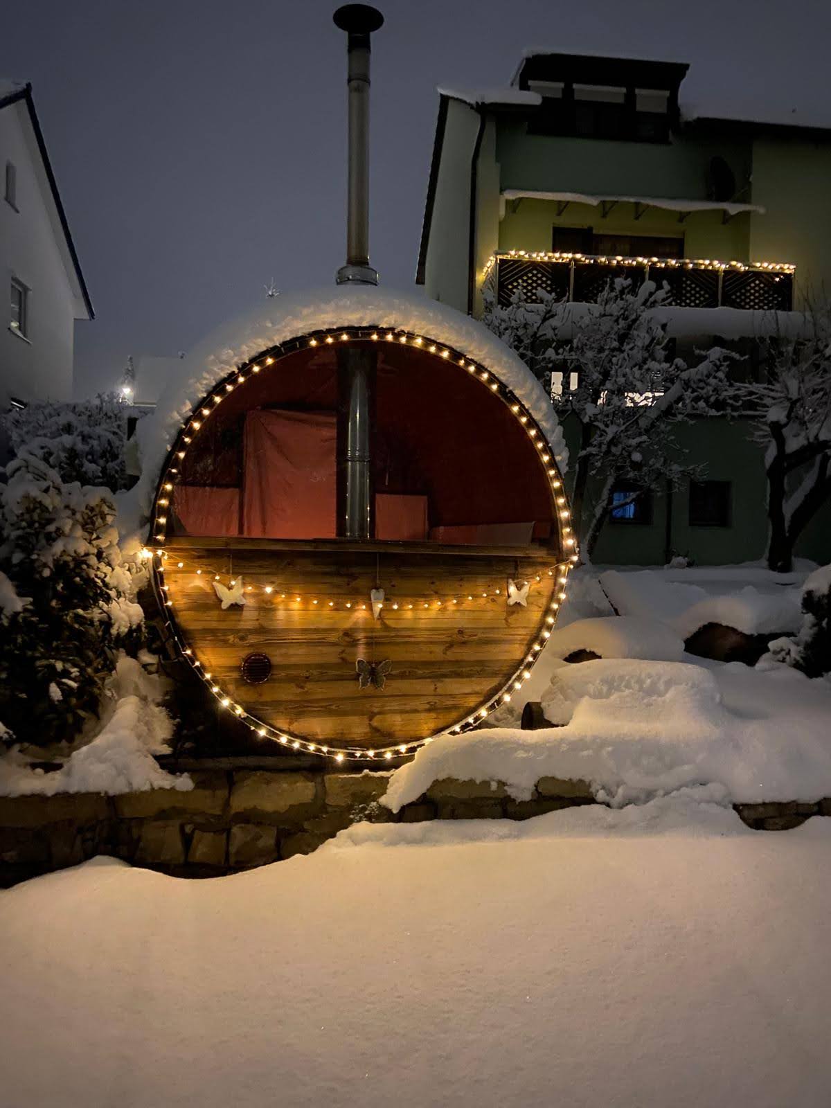 Fasssauna mit Schornstein und Lichterkette in einem verschneiten Garten