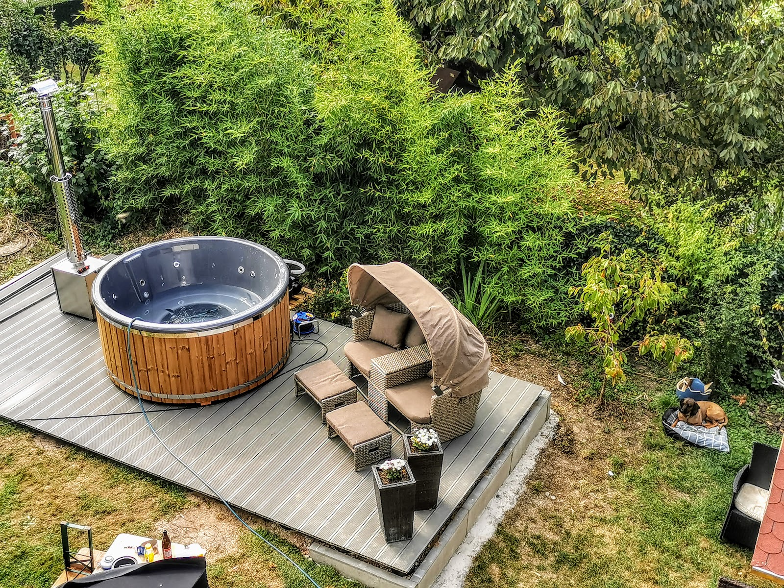 Filteranlage 1 Rundes Badefass mit dunkelgrauer Innenwanne und klarem Wasser auf einer Holzterrasse, daneben Außenofen mit Edelstahlschornstein und Gartenschlauch zum Befüllen