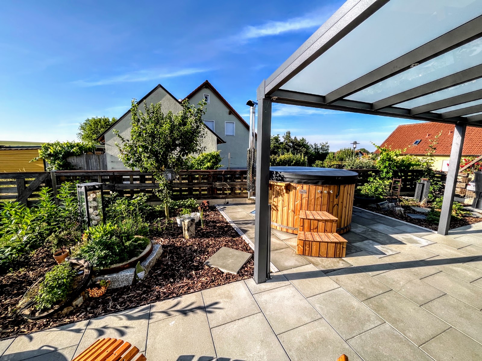 Reinigung 1 Rundes Holzbadefass mit dunkler Abdeckung und Außenofen mit Edelstahlschornstein auf einer gefliesten Terrasse unter einer Glaspergola