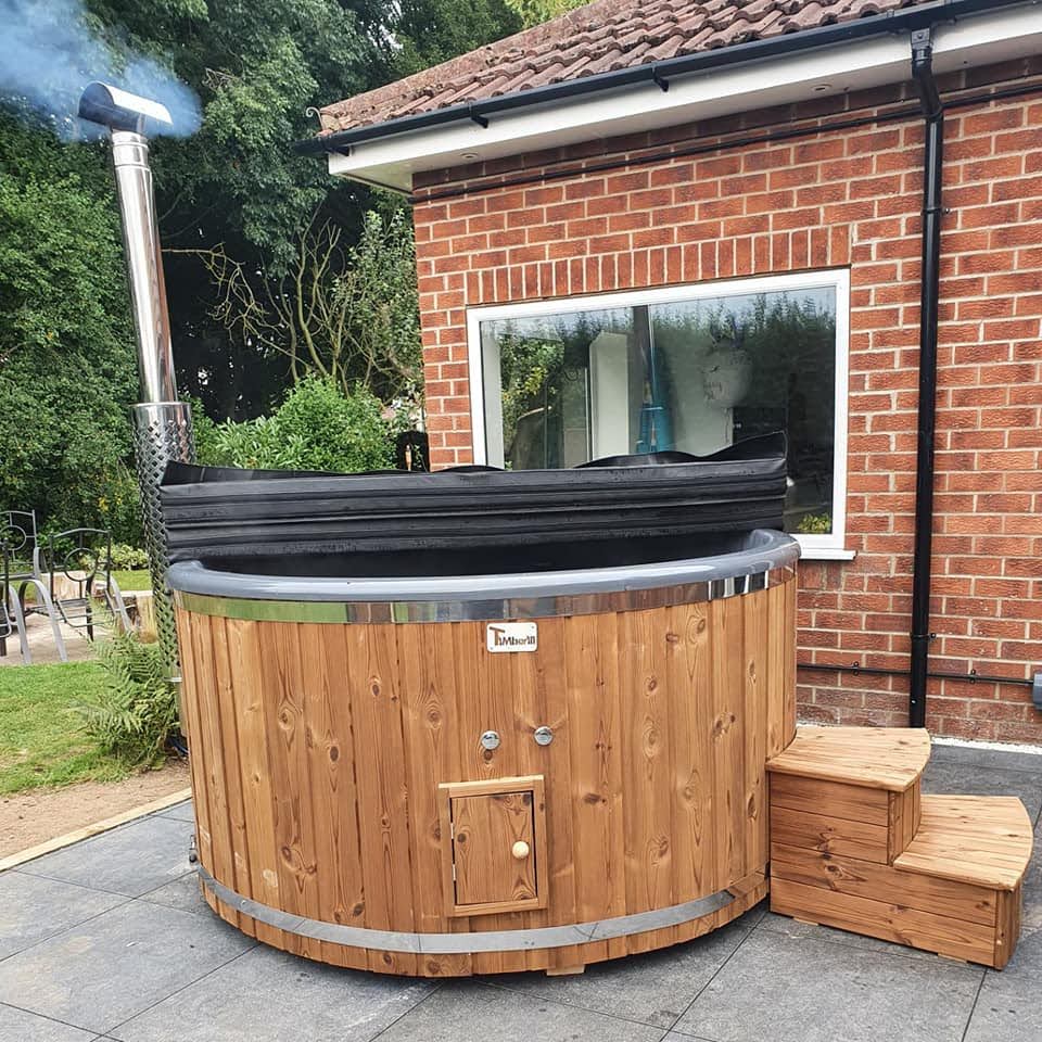 Runder Hot Tub aus Holz mit Metallbändern, Außenofen mit Edelstahlschornstein, zusammengeklappter schwarzer Abdeckung und Holztreppe auf einer Terrasse vor einem Backsteinhaus