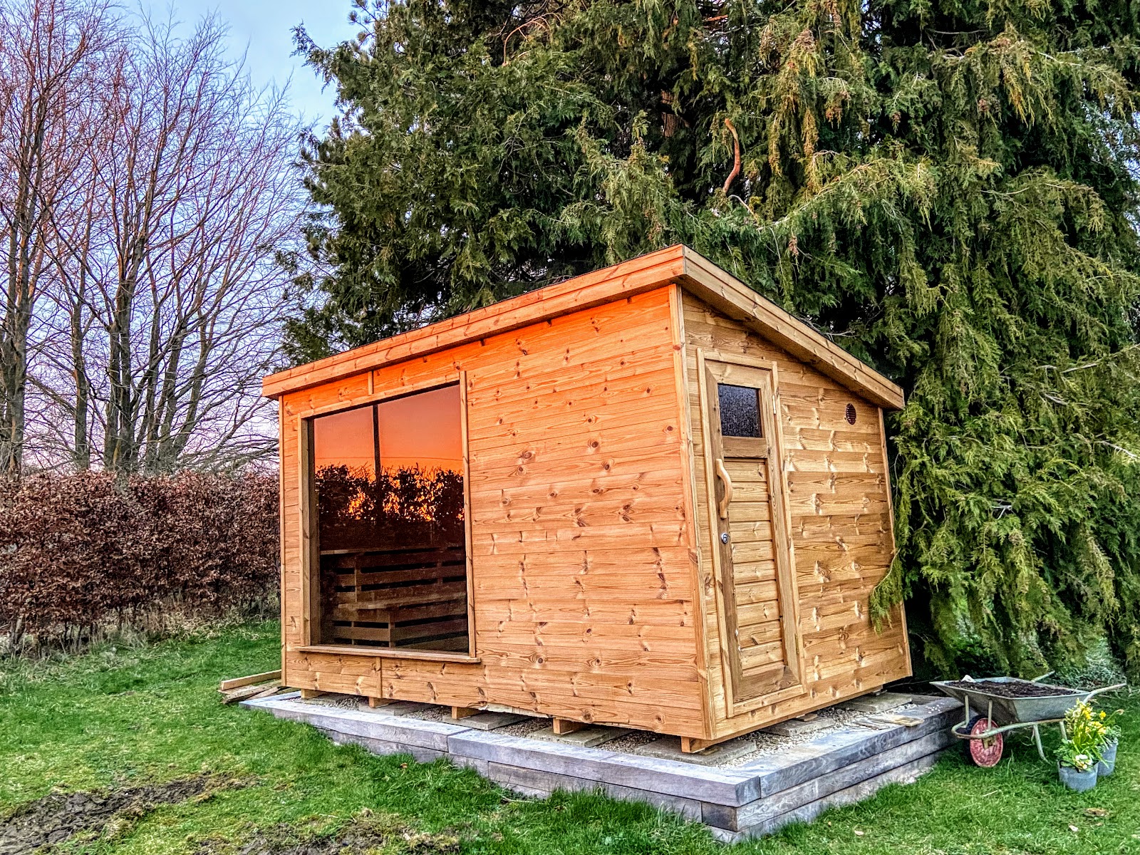 Rechteckiges Saunahaus mit flach geneigtem Dach, großer Glasfront und honigbrauner Holzverkleidung auf einem Holzpodest im Garten