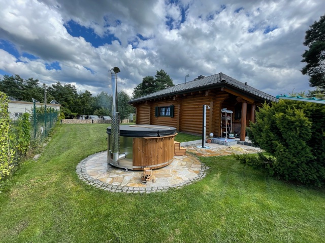 Aufstellen 1 Rundes Holzbadefass mit schwarzer Abdeckung, kleiner Holzstufe und Außenofen mit Edelstahlschornstein auf einer runden Pflasterfläche mit Randsteinen im Rasen vor einem Blockhaus
