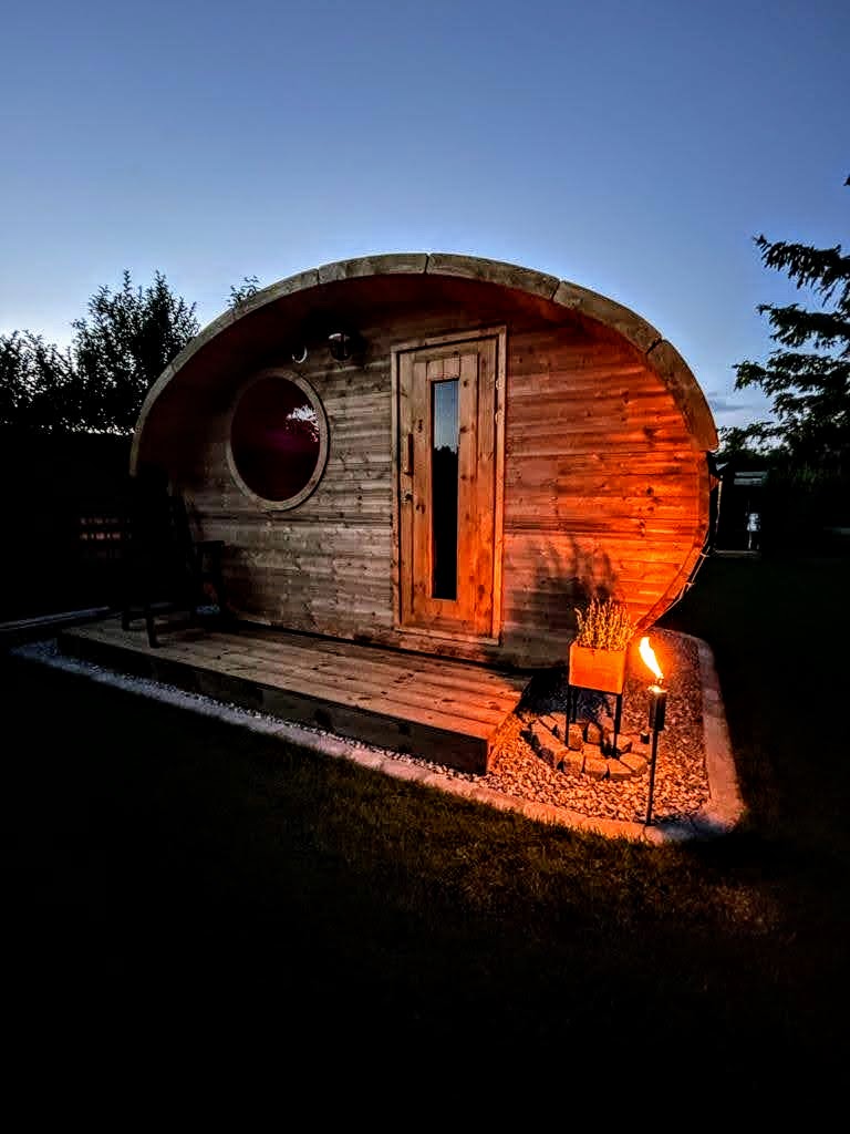Kleine Fasssauna aus Holz mit Rundfenster, abends im Garten beleuchtet