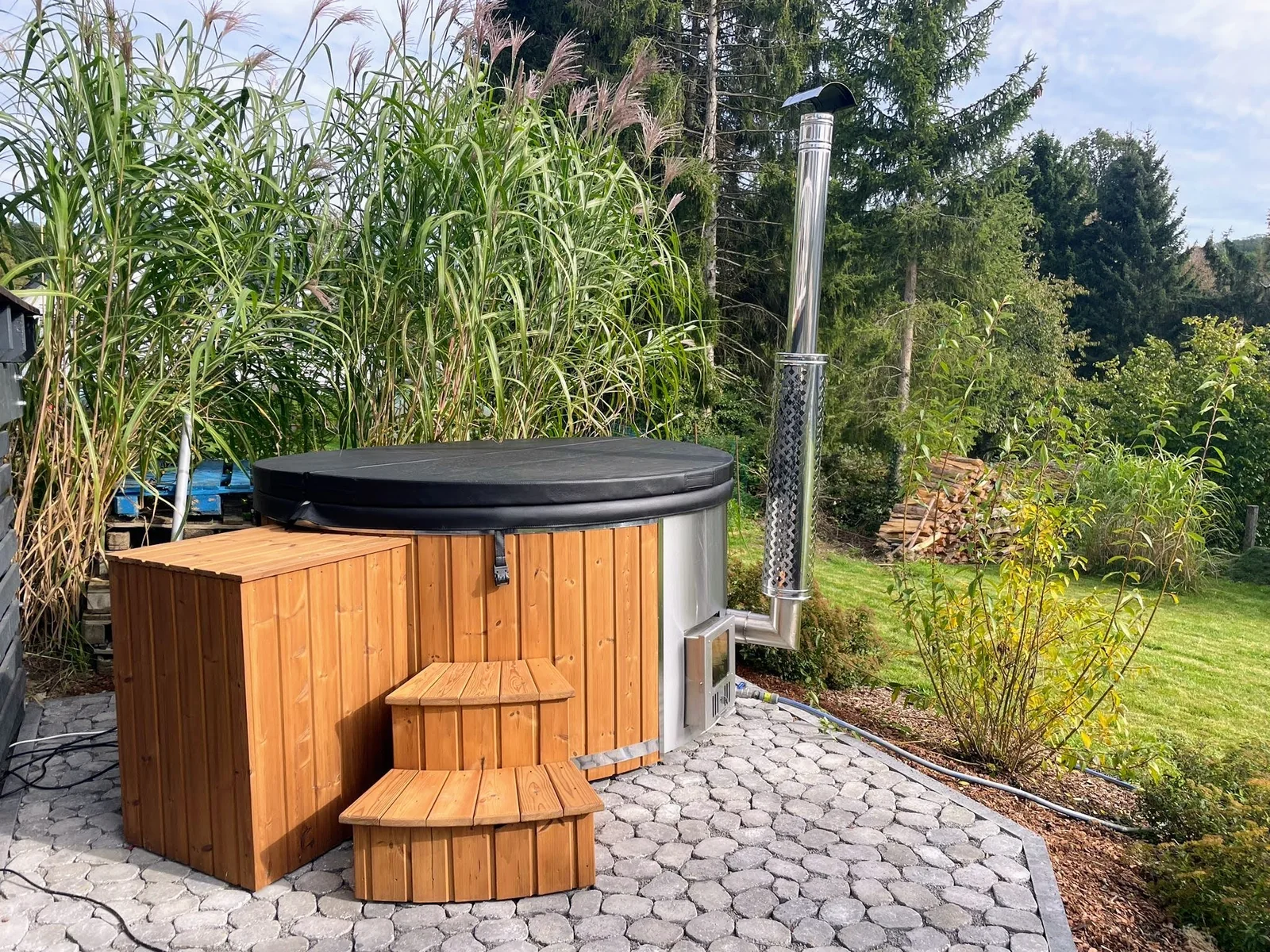 kaufen 1 Badefass mit Holzverkleidung, schwarzer Abdeckung, Holztreppe und Ofen mit Schornstein auf einer gepflasterten Gartenfläche.