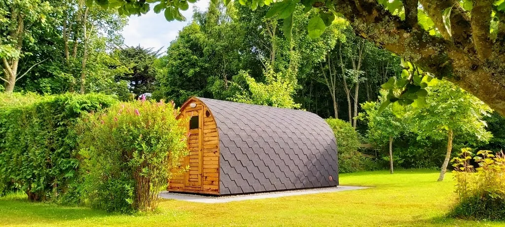 klein 1 Kompakte bogenförmige Außensauna mit Holztür und dunklem Schindeldach in einem grünen Garten.